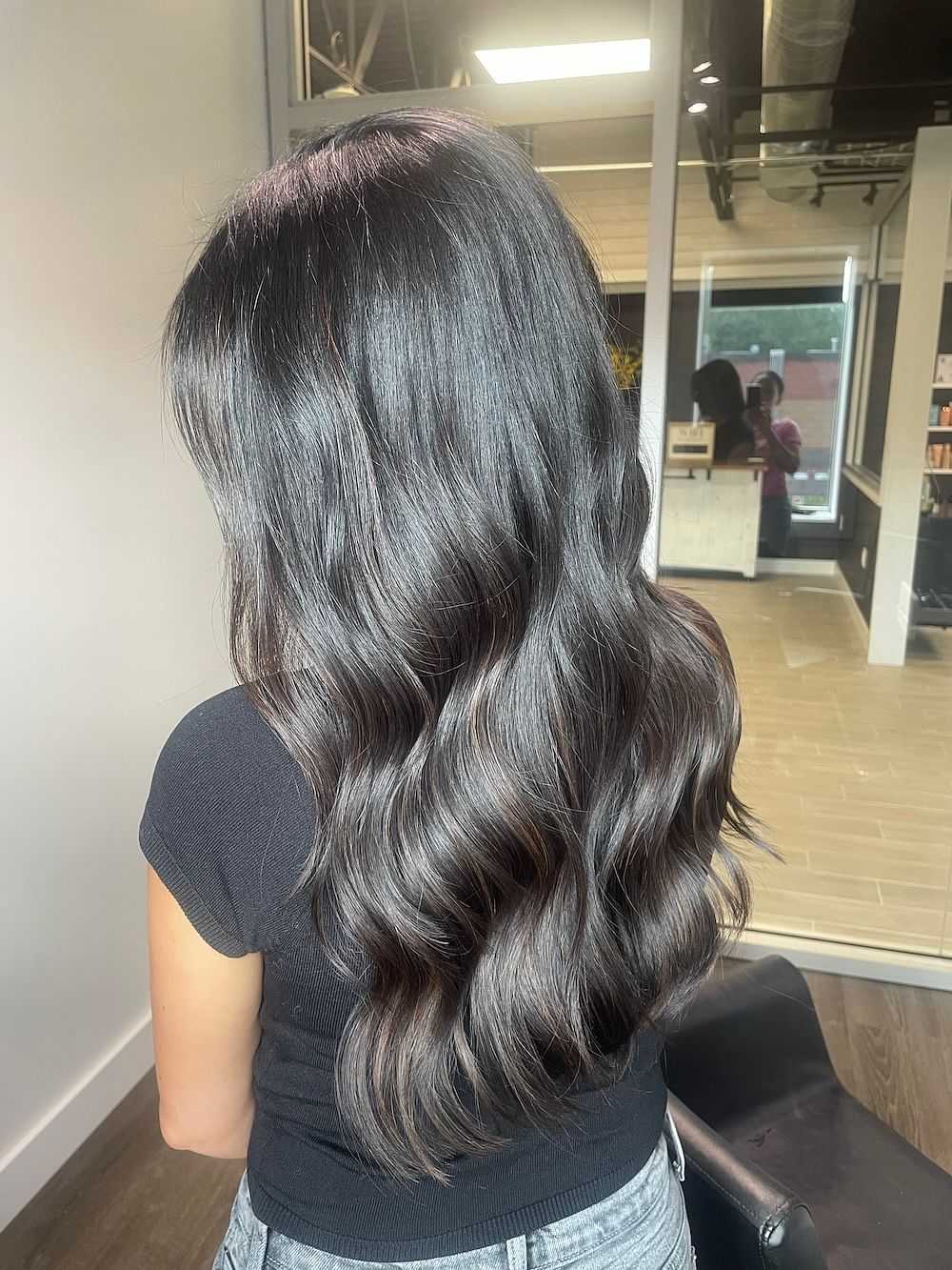 Woman with long, wavy, dark hair in a salon, facing away from the camera.